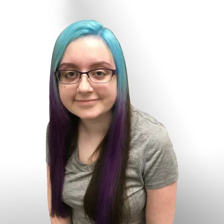 An image of a young woman with glasses against a white backdrop. Her skin is caucasian, she is wearing a grey striped shirt, and her hair is long, straight, and has bangs in a gradient of bright cyan on top to purple at the ends, the rest of her hair being a dark brown.